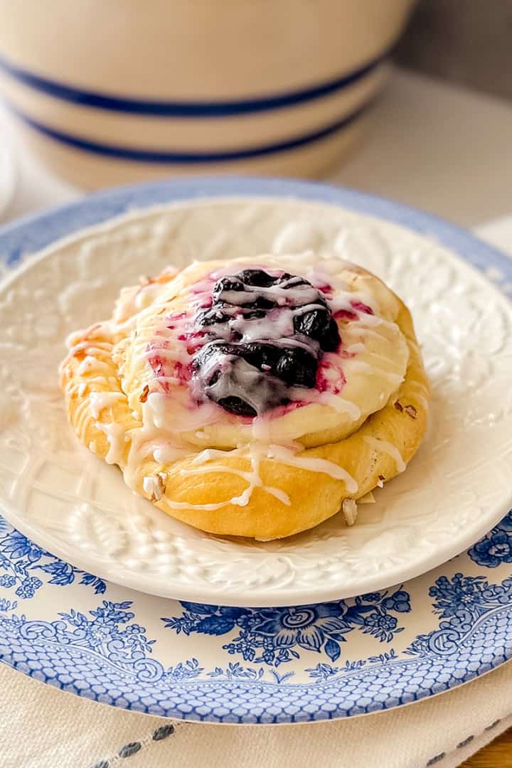 A Danish bun topped with white icing and a luscious blueberry filling sits on a white plate with a blue floral border, placed on a white napkin.
