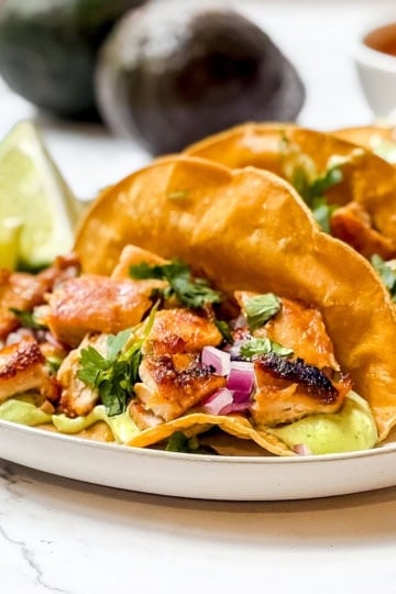 Three easy chicken tacos on a plate with lime wedges and avocados in the background.