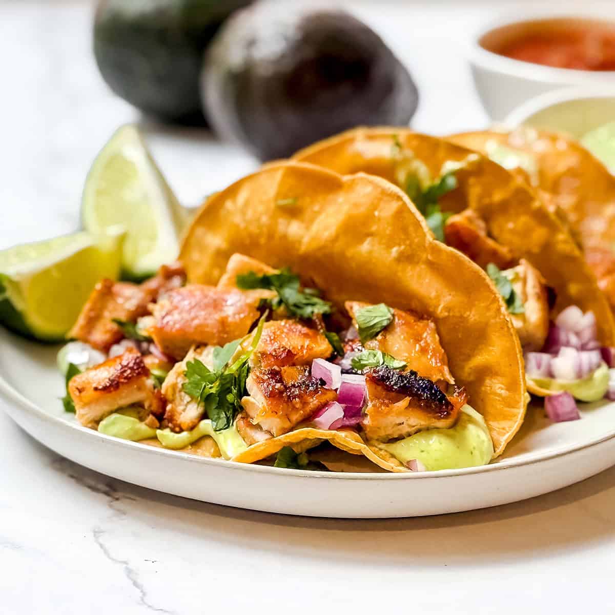 Three easy chicken tacos on a plate with lime wedges and avocados in the background.
