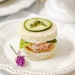 A round tea sandwich with a ham salad filling, leafy greens, a cucumber slice, and dill on top is served on a white plate with a small purple flower garnish—an elegant addition to your collection of easy recipes for tea sandwiches.