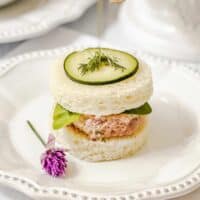 A round tea sandwich with a ham salad filling, leafy greens, a cucumber slice, and dill on top is served on a white plate with a small purple flower garnish-an elegant addition to your collection of easy recipes for tea sandwiches.