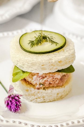 A round tea sandwich with a ham salad filling, leafy greens, a cucumber slice, and dill on top is served on a white plate with a small purple flower garnish-an elegant addition to your collection of easy recipes for tea sandwiches.