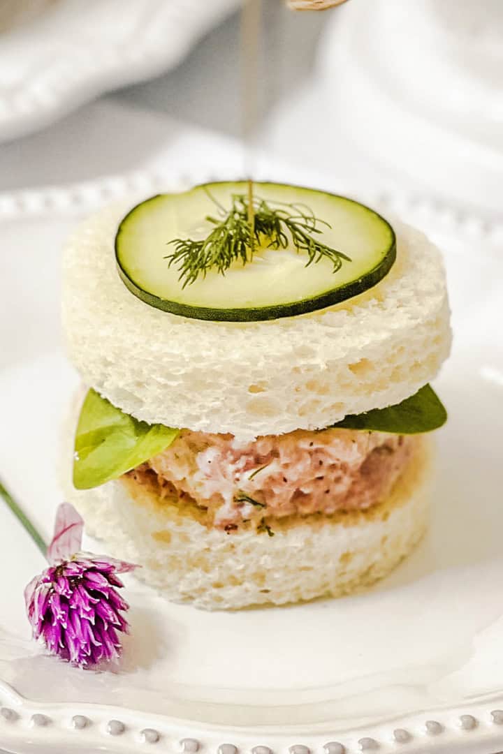 A round tea sandwich with a ham salad filling, leafy greens, a cucumber slice, and dill on top is served on a white plate with a small purple flower garnish-an elegant addition to your collection of easy recipes for tea sandwiches.