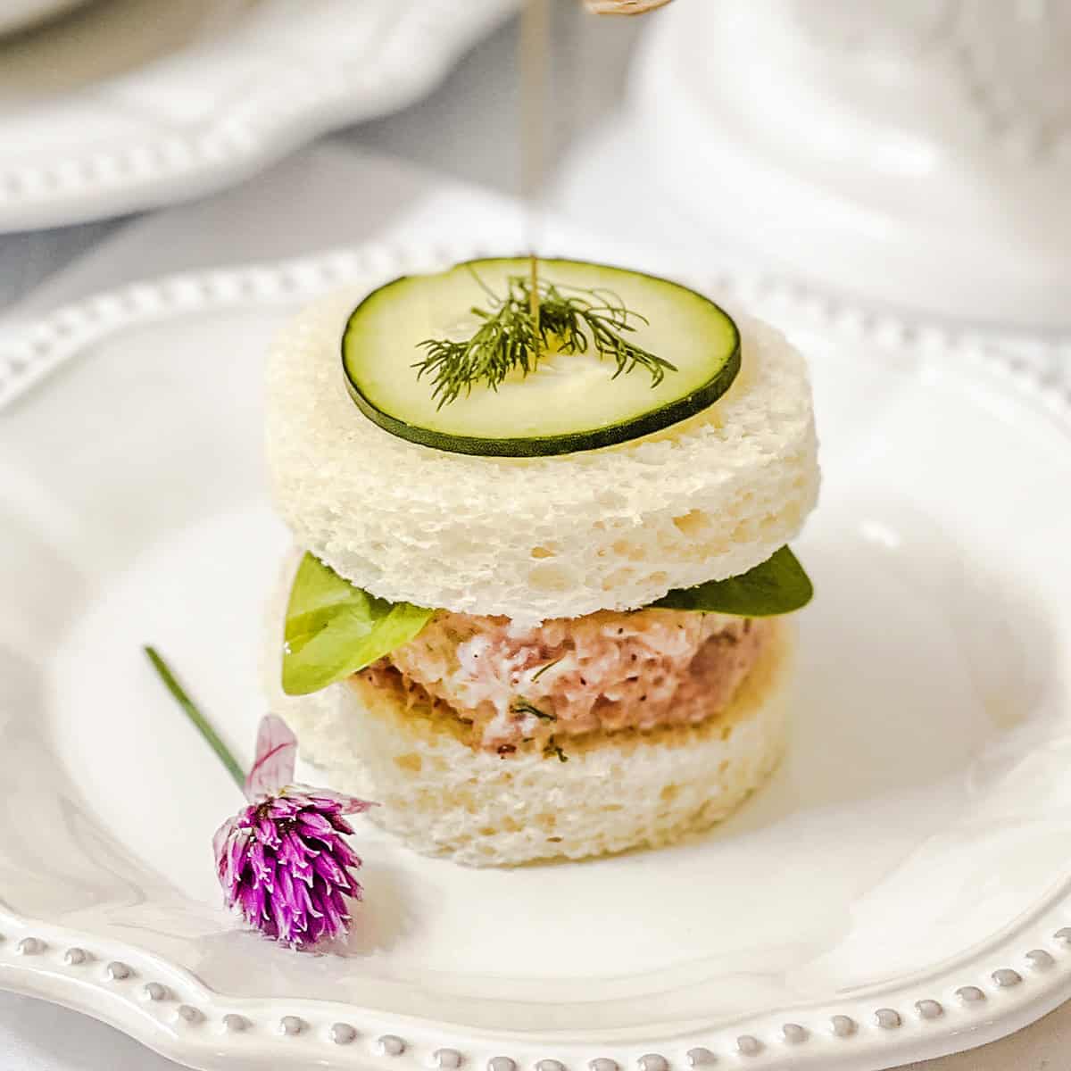 A round tea sandwich with a ham salad filling, leafy greens, a cucumber slice, and dill on top is served on a white plate with a small purple flower garnish-an elegant addition to your collection of easy recipes for tea sandwiches.