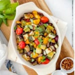 White oval dish filled with roasted ratatouille including tomatoes, zucchini, and eggplant, garnished with fresh herbs.