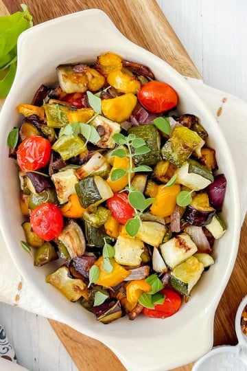 White oval dish filled with roasted ratatouille including tomatoes, zucchini, and eggplant, garnished with fresh herbs.