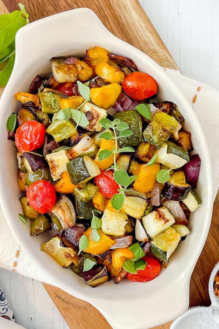 White oval dish filled with roasted ratatouille including tomatoes, zucchini, and eggplant, garnished with fresh herbs.