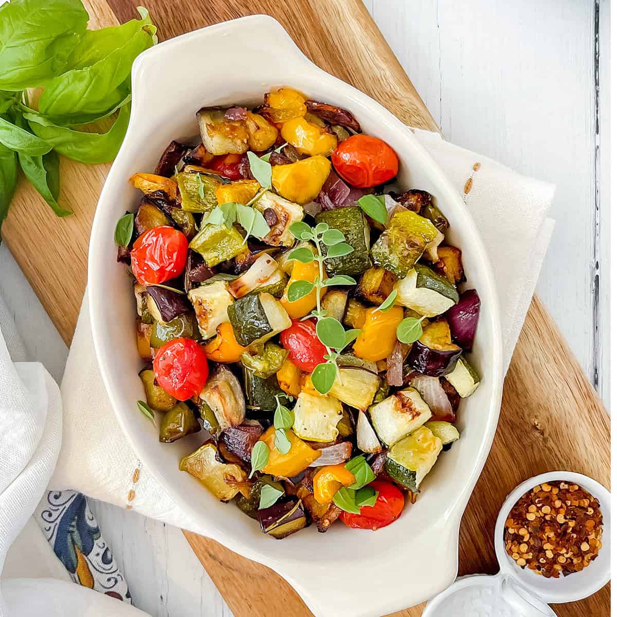 White oval dish filled with roasted ratatouille including tomatoes, zucchini, and eggplant, garnished with fresh herbs.