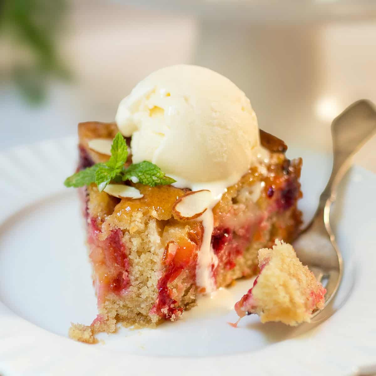 Slice of plum almond cake topped with vanilla ice cream and mint, showing fresh baked plums and a tender almond crumb with a fork nearby.