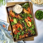 Sheet pan chicken fajitas with seasoned chicken, black beans, kale, red peppers, and lime wedges. A bowl of creamy sauce sits on the pan.