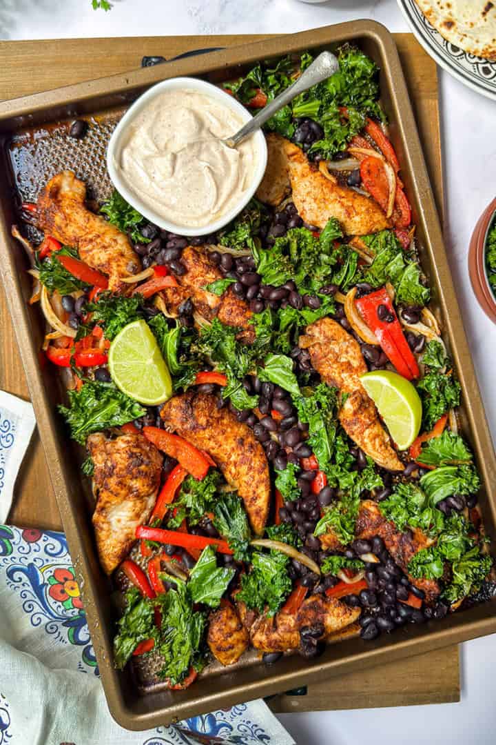 Sheet pan chicken fajitas with seasoned chicken, black beans, kale, red peppers, and lime wedges. A bowl of creamy sauce sits on the pan.