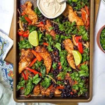 Closeup view of sheet pan chicken fajitas with peppers, kale, black beans, and lime wedges.