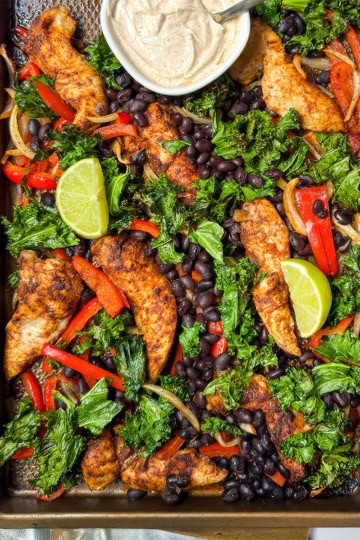 Closeup view of sheet pan chicken fajitas with peppers, kale, black beans, and lime wedges.