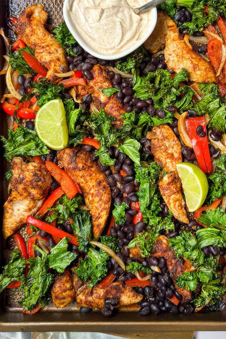 Closeup view of sheet pan chicken fajitas with peppers, kale, black beans, and lime wedges.