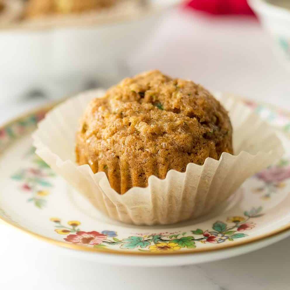 A single muffin in a paper liner sits on a floral-patterned plate, with more muffins and a teacup blurred in the background, creating a cozy scene perfect for enjoying freshly baked muffins.
