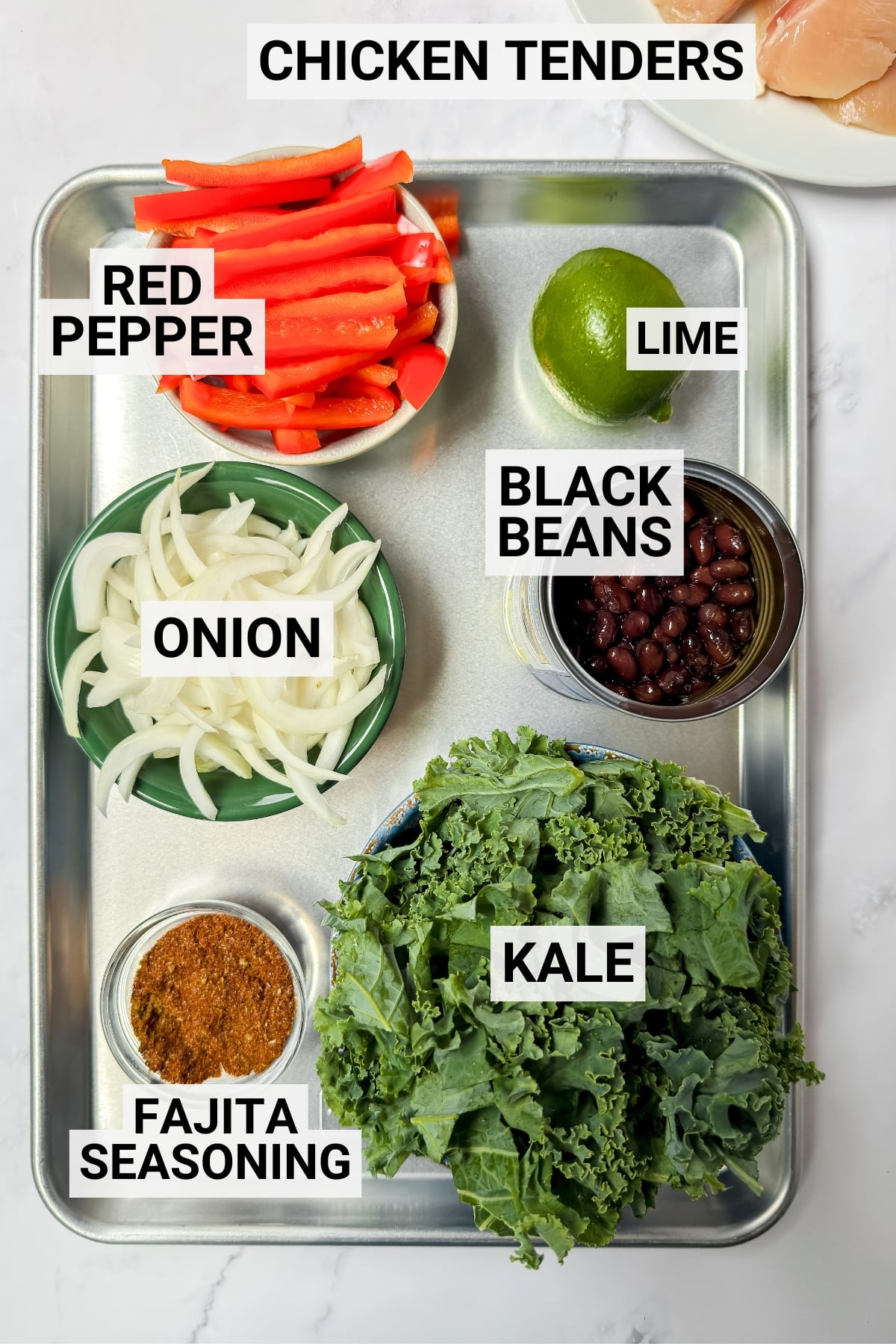 A metal tray holds all the components for sheet pan chicken fajitas: raw chicken tenders, sliced red pepper, lime, black beans, sliced onion, fajita seasoning, and kale-each ingredient clearly labeled with text.