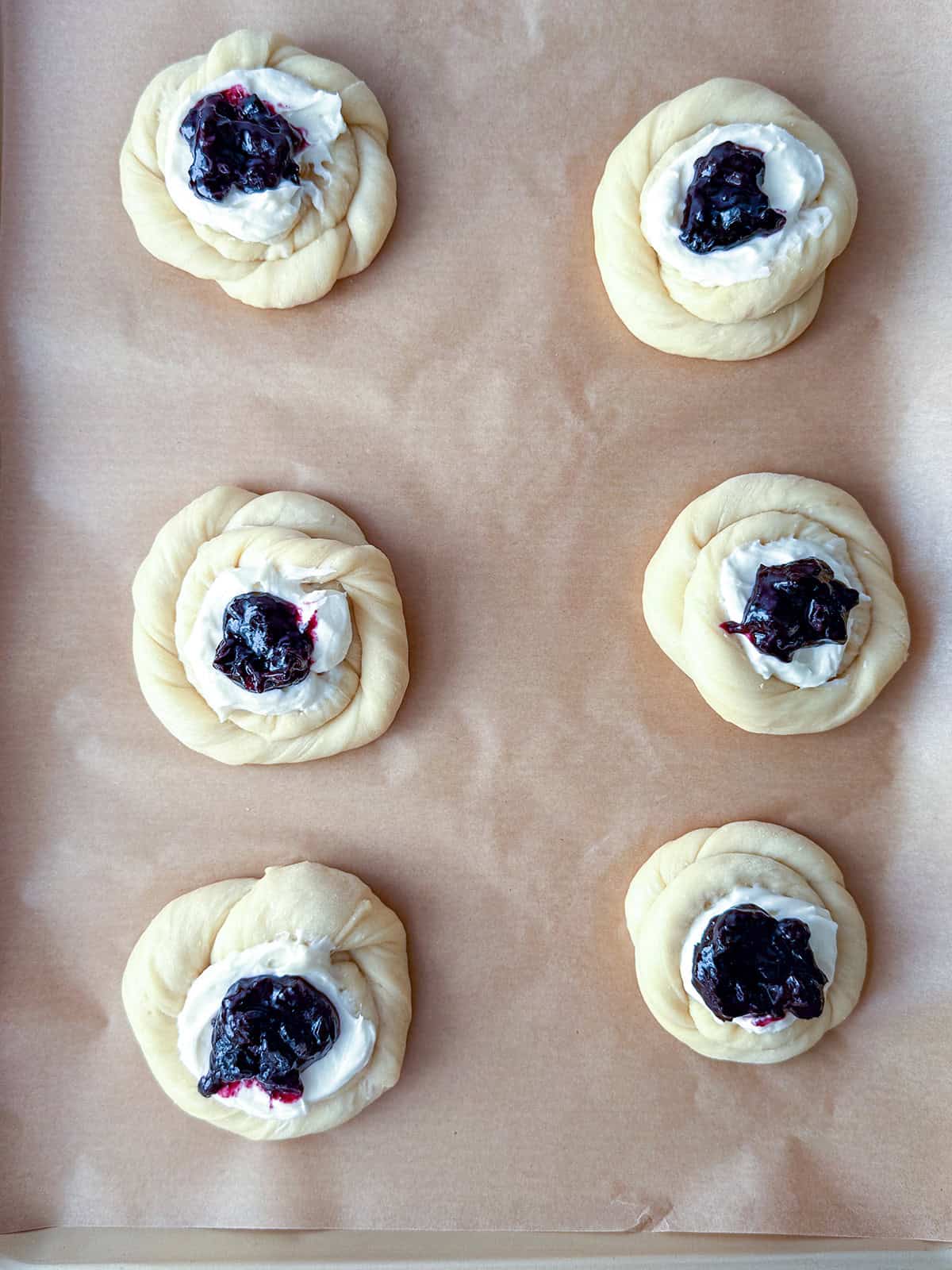 Six unbaked Blueberry Danish pastries with cream cheese and fruit filling, made from yeasted sweet dough, are arranged in two rows of three on a parchment-lined baking sheet.