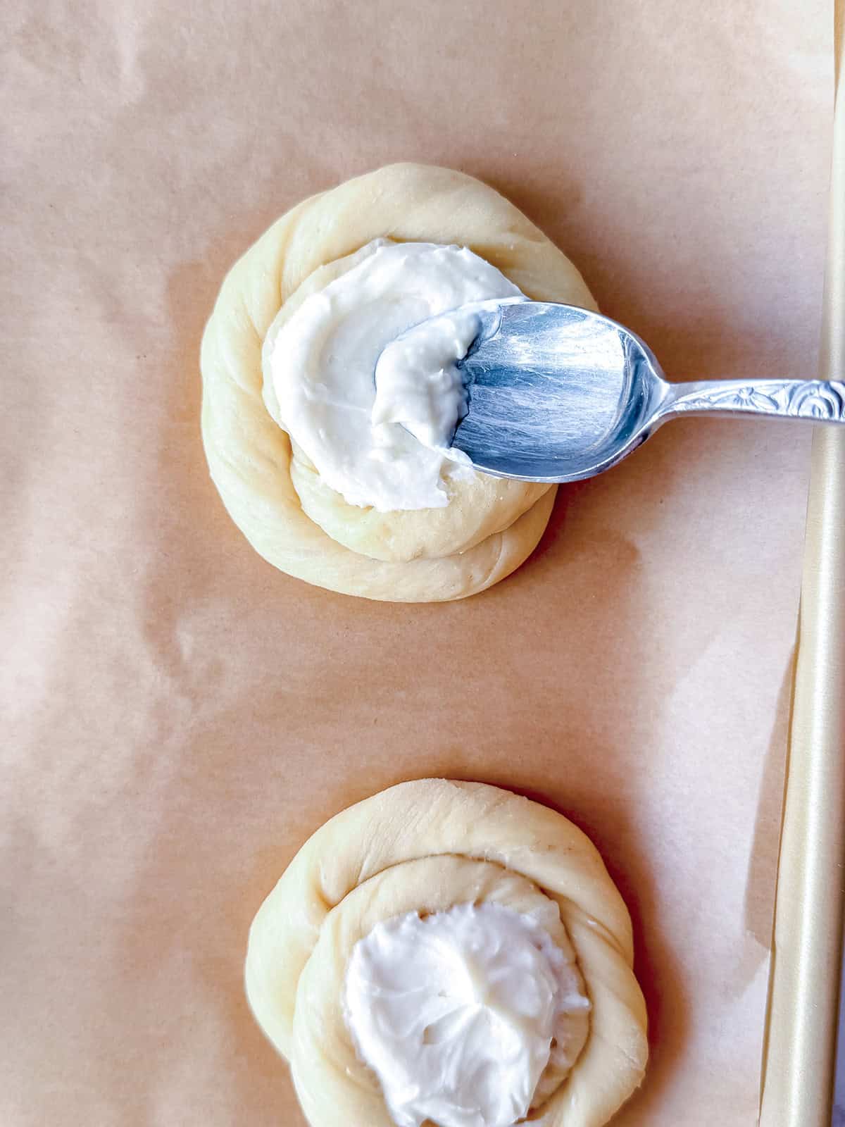 A spoon spreads creamy filling onto a round, twisted Yeasted Sweet Dough placed on parchment paper, with another filled dough below it-perfect for making homemade Blueberry Danish pastries.