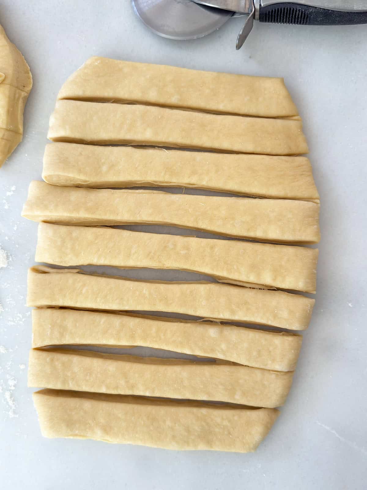 Rectangular sheet of Sweet Dough cut into even strips, with a pastry cutter and fork visible at the top right corner on a white surface-perfect for making a classic Blueberry Danish.