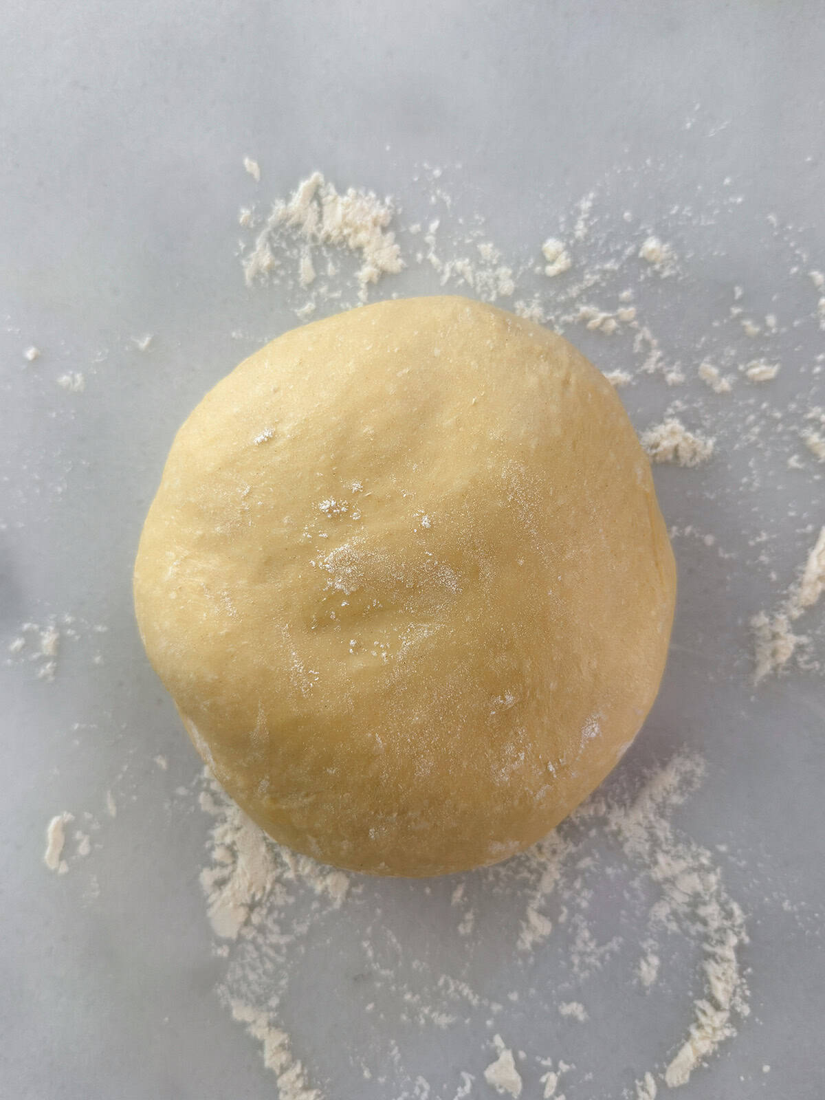 A round ball of yeasted sweet dough rests on a lightly floured surface, with scattered flour visible around it.