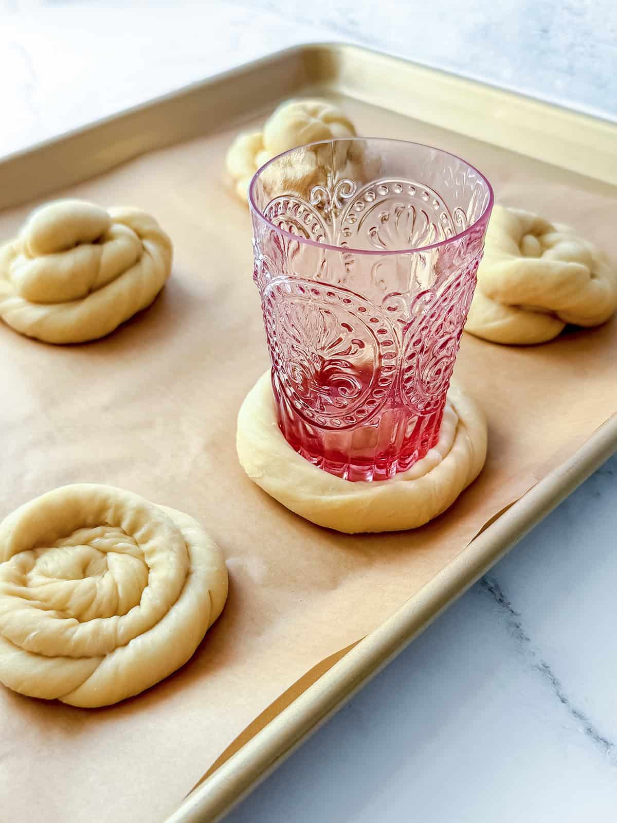 A decorative pink glass presses into a ring of sweet dough on a baking sheet lined with parchment paper; other unshaped dough rings, ready to become Blueberry Danish, are nearby.