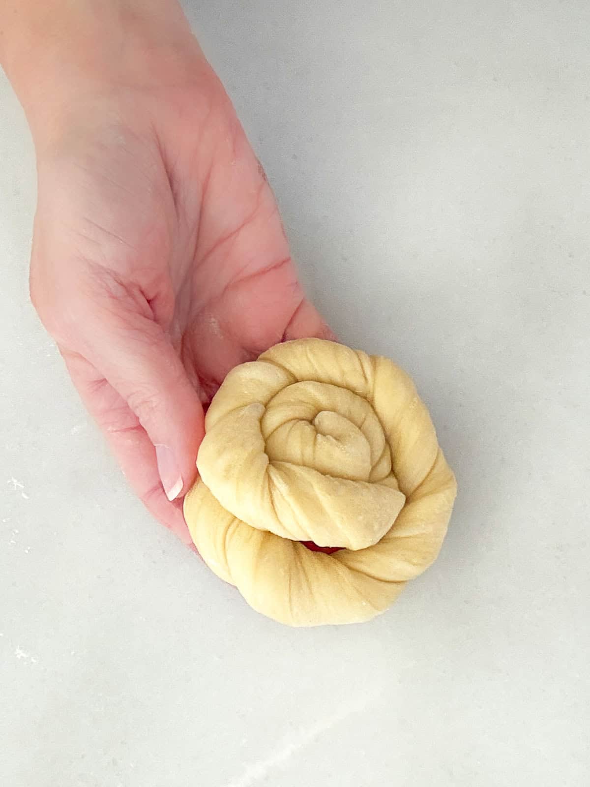 A hand holding a twisted, unbaked yeasted sweet dough shaped into a spiral on a light surface.