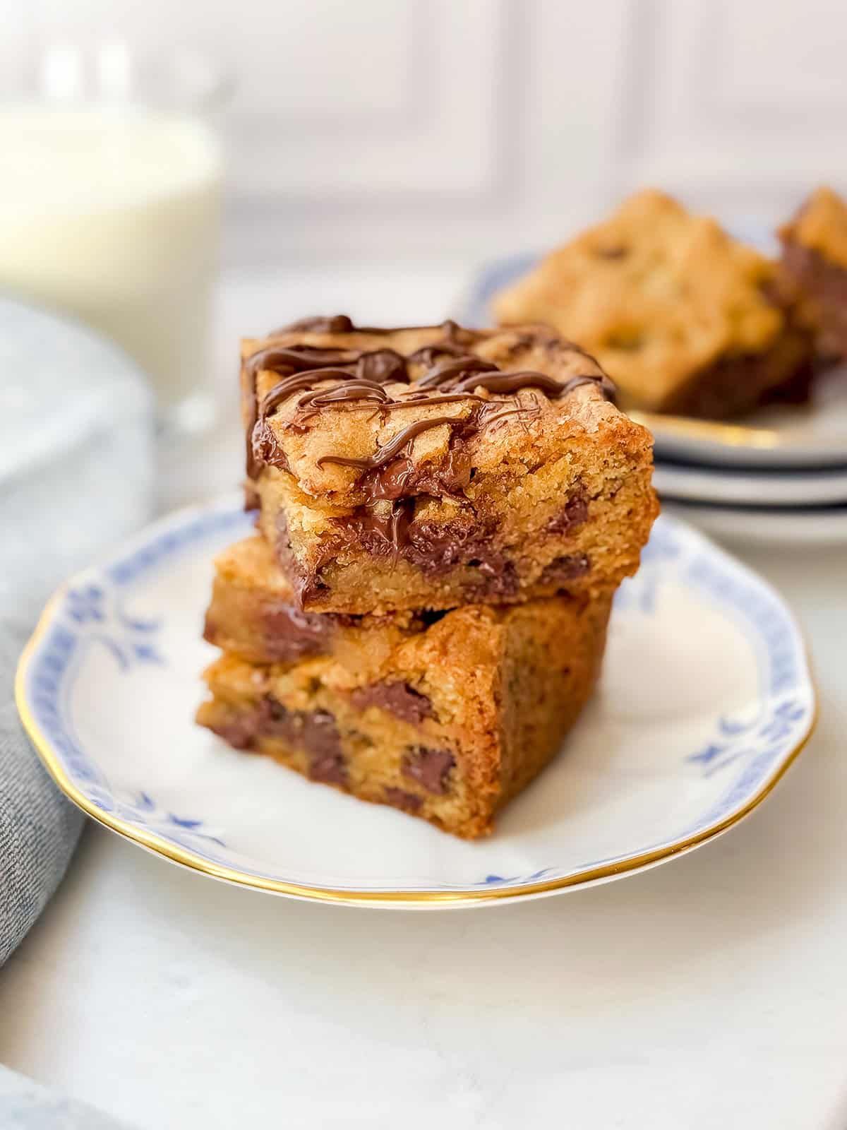 Stacked chocolate chip cookie bars drizzled in chocolate with more on a plate in the background.