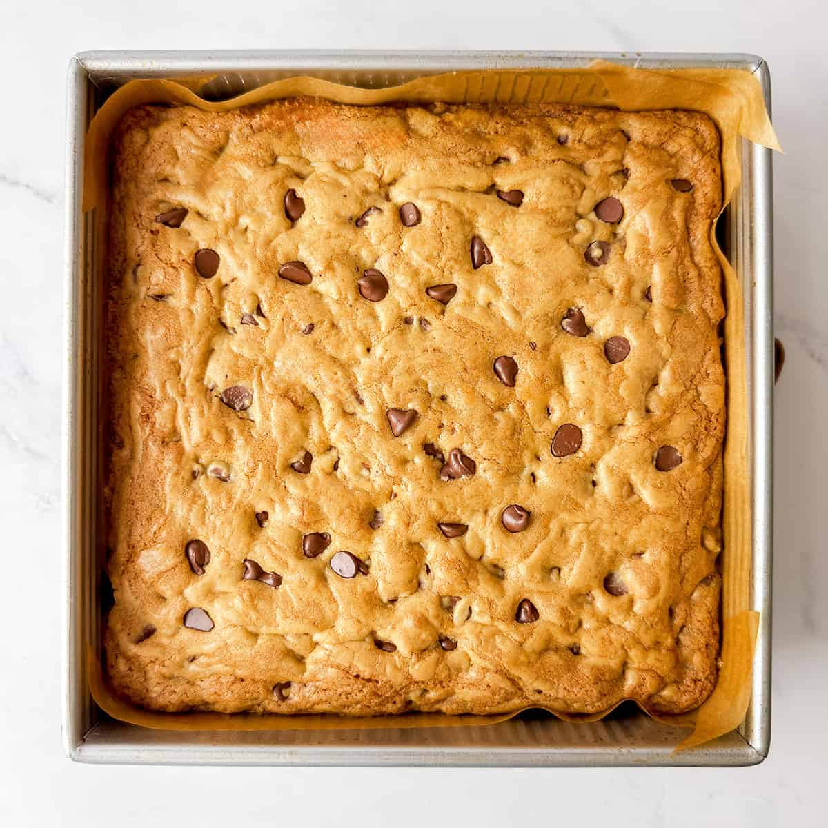 Baked chocolate chip cookie bars cooling in a parchment lined square baking pan on a marble counter.