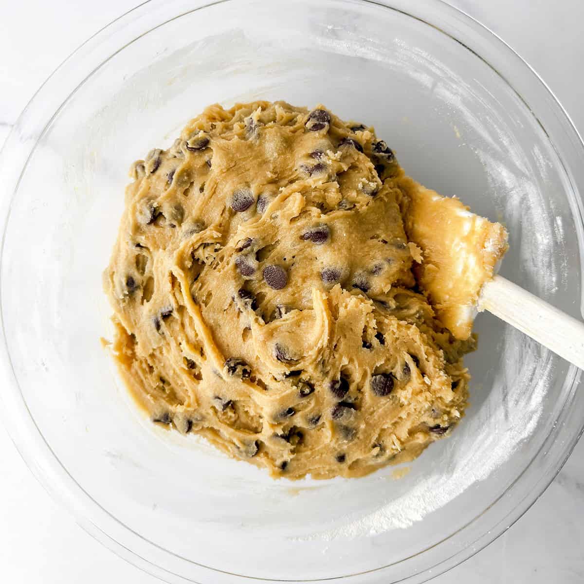 Chocolate chip cookie batter mixed together in a glass bowl with a rubber spatula.