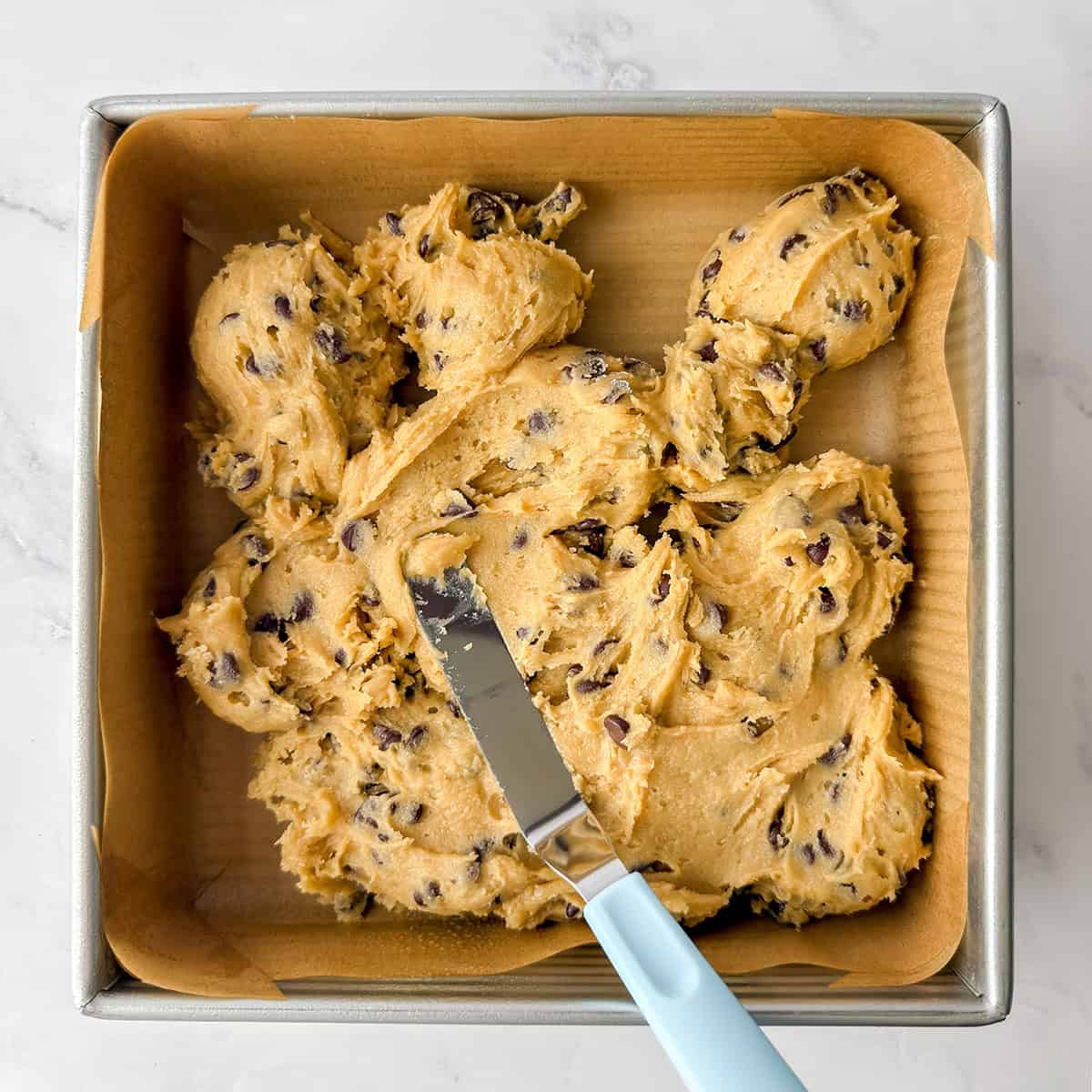A parchment lined square baking pan with dollops of cookie batter and an offset spatula for spreading.