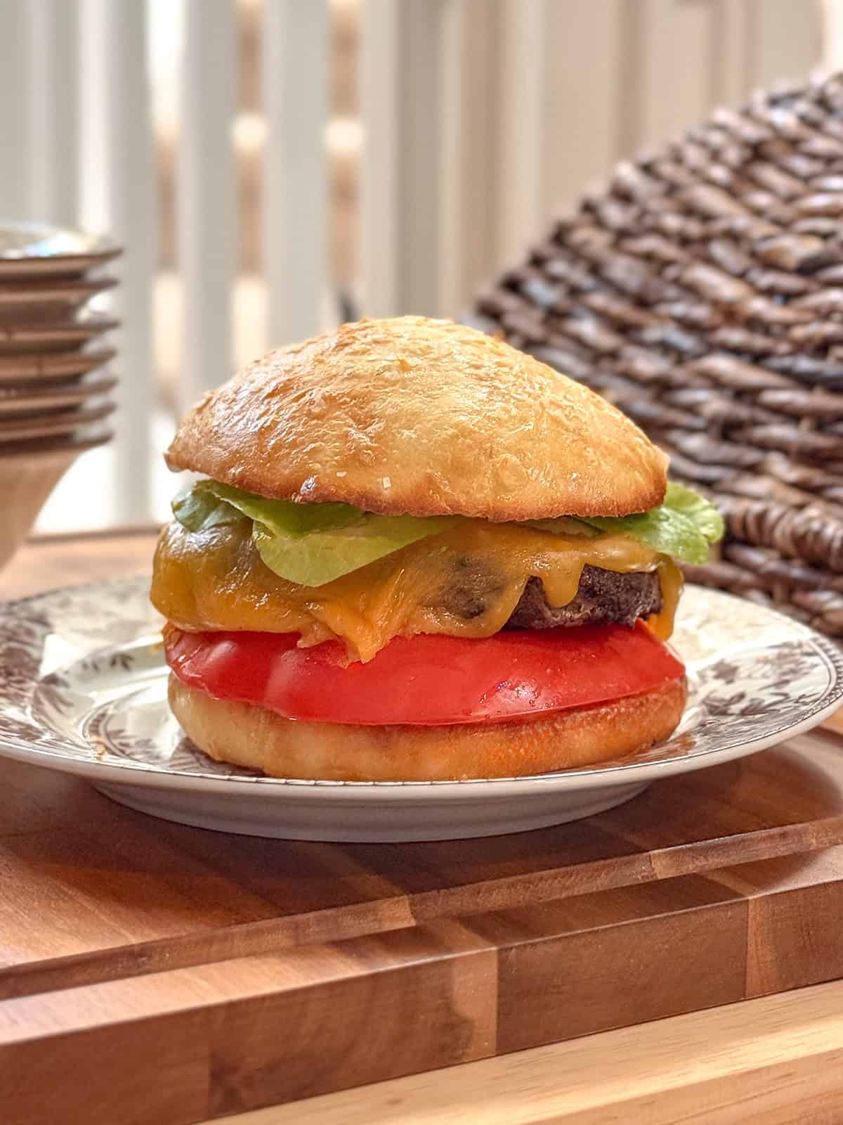 Grilled hamburger on a wooden serving board with a homemade hamburger bun, melted cheddar cheese, lettuce, and tomato.