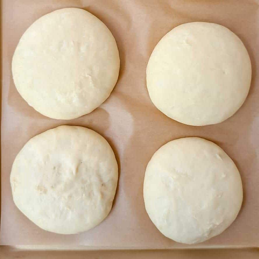 Eight shaped hamburger buns rising on a parchment-lined baking sheet.