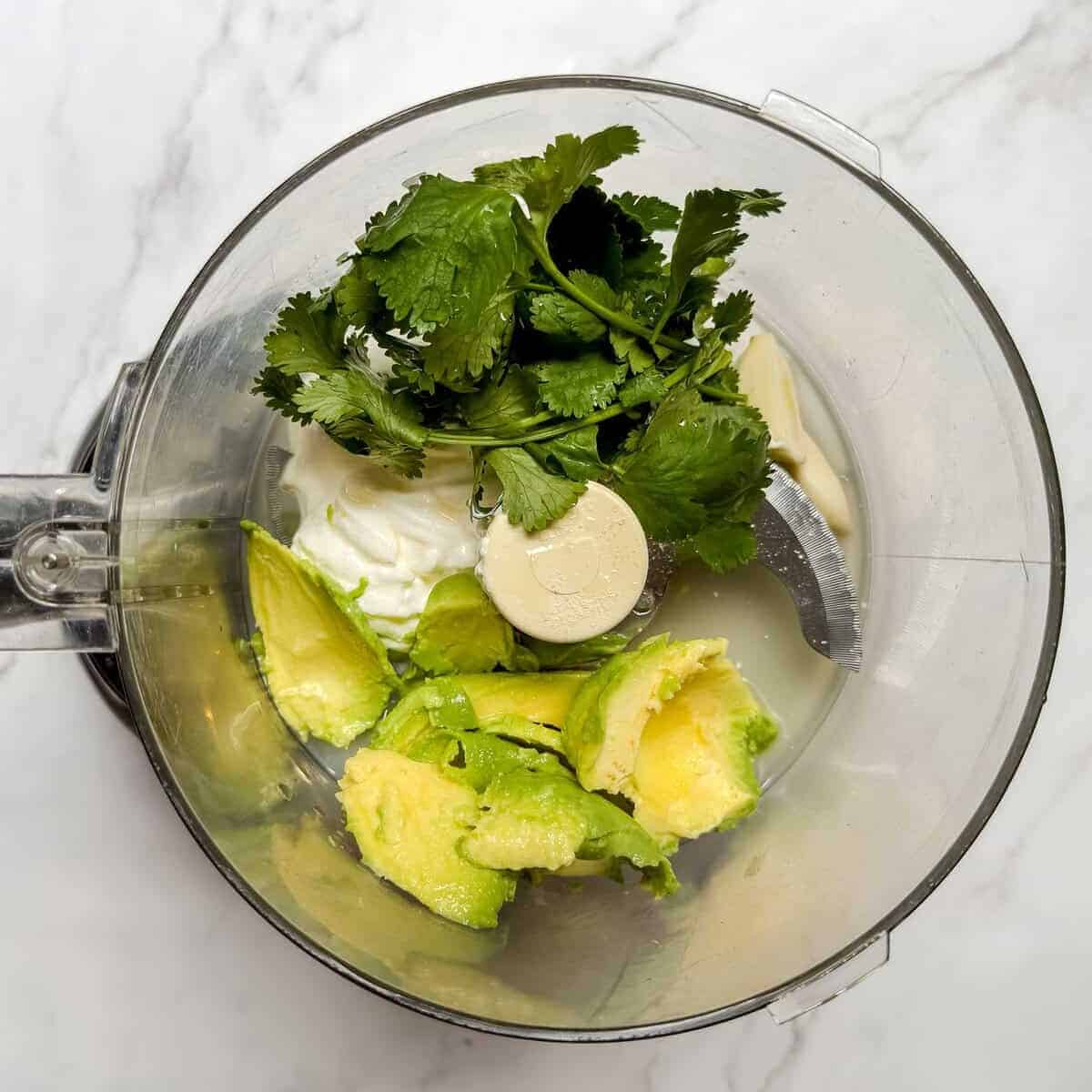 A food processor bowl containing avocado slices, fresh cilantro, and a plain Greek yogurt sits on a marble countertop, perfectly prepped for delicious chicken tacos.