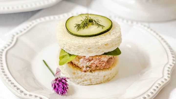 A round tea sandwich with white bread, salmon filling, spinach leaves, cucumber slice, and dill, served on a white plate with a small purple flower—perfect for easy recipes or your next afternoon gathering.