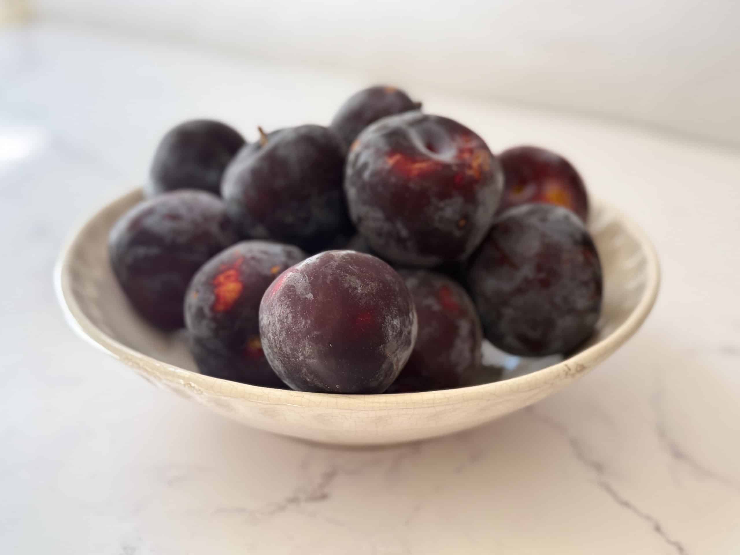 Bowl of fresh plums sitting on a marble counter.
