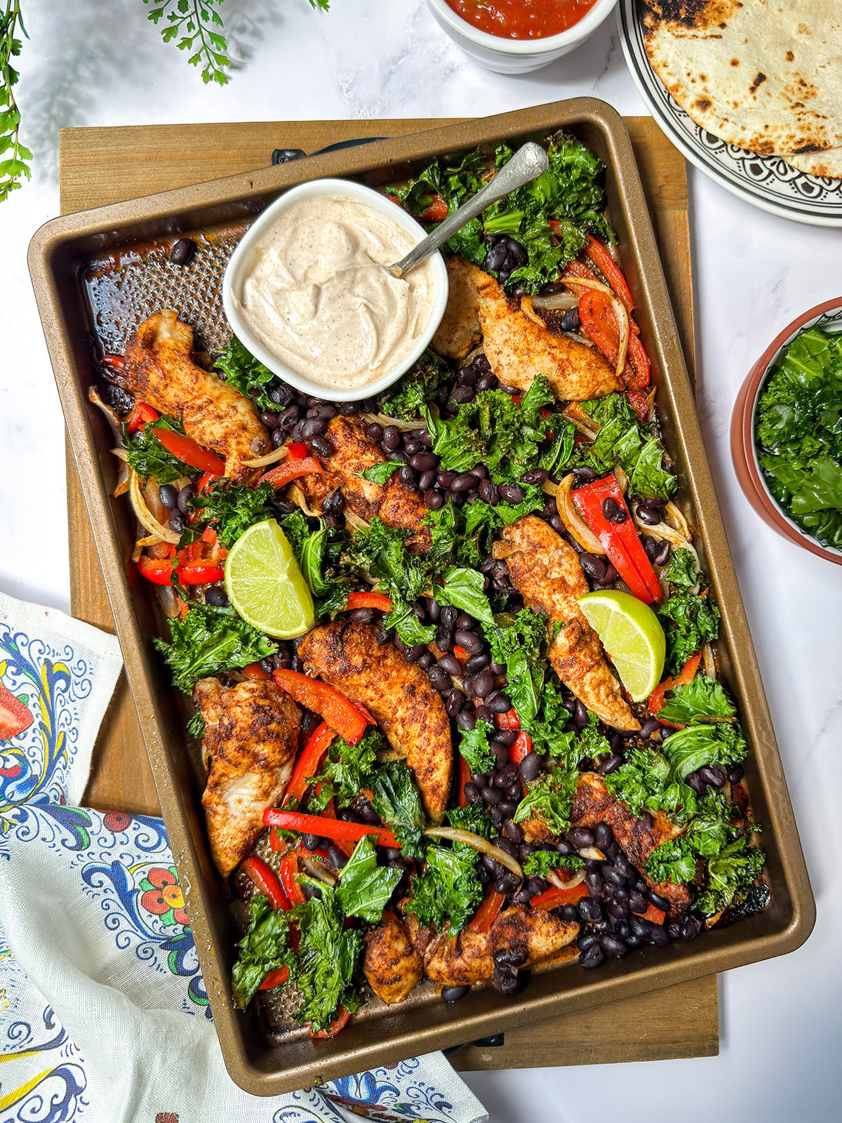 Sheet pan chicken fajitas with seasoned chicken strips, black beans, kale, red peppers, onions, lime wedges, and a bowl of creamy sauce. Tortillas and salsa are visible nearby.