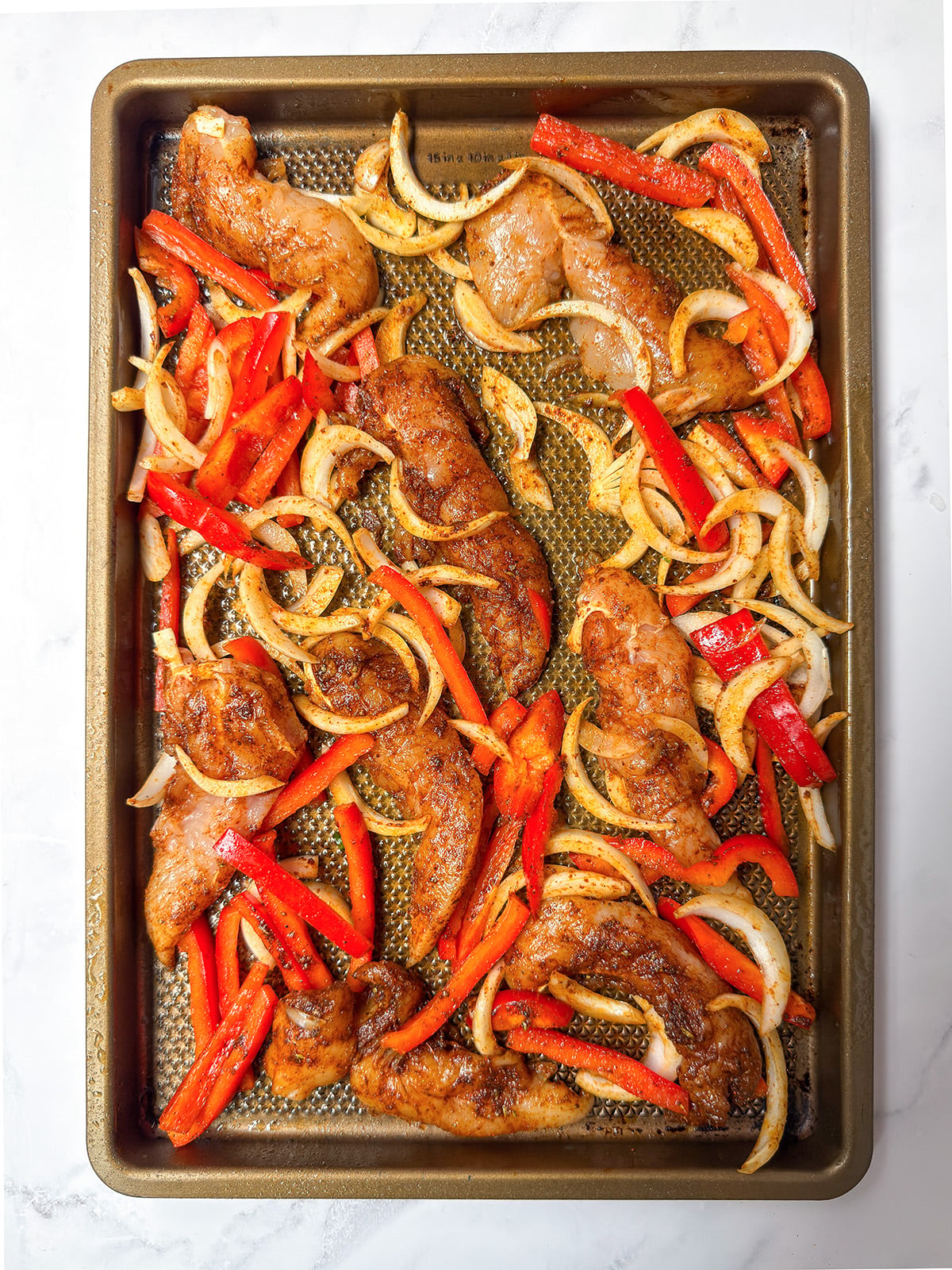 Sheet pan chicken fajitas feature seasoned chicken strips, sliced red bell peppers, and onions arranged evenly, ready for baking.
