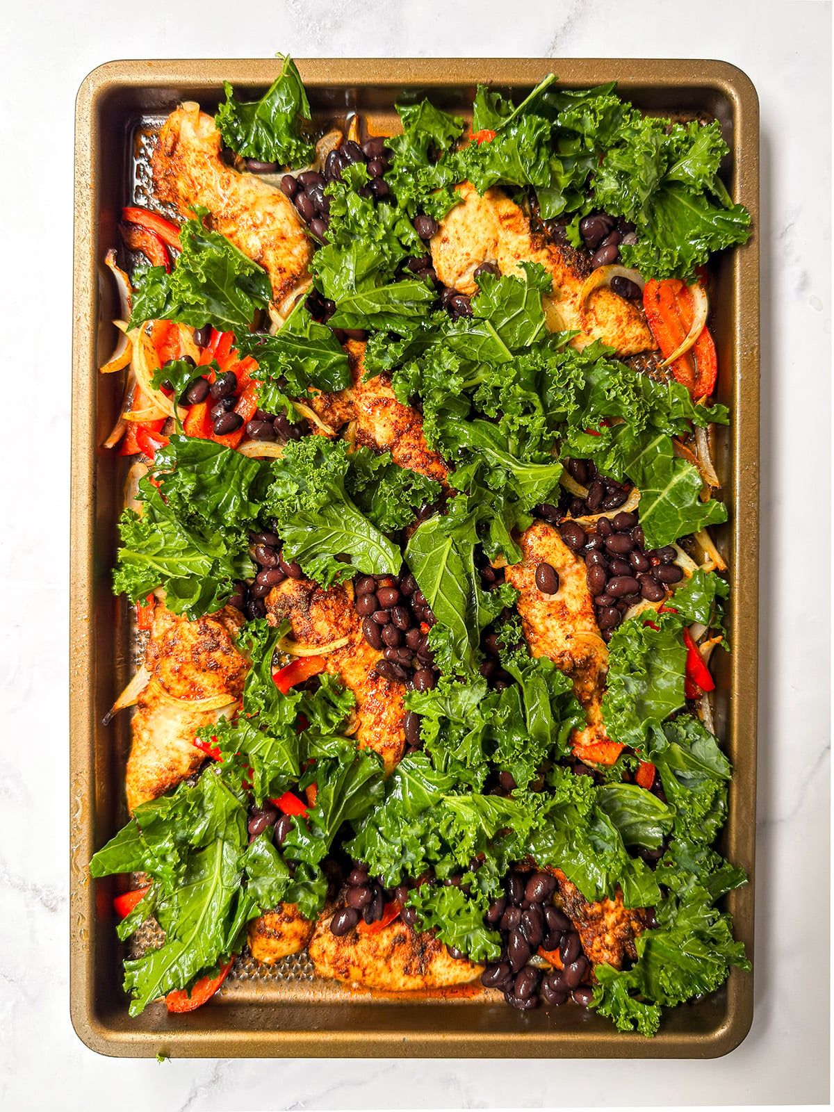 Sheet pan chicken fajitas featuring baked seasoned chicken, black beans, kale, sliced onions, and red bell peppers are arranged on a metal baking tray.