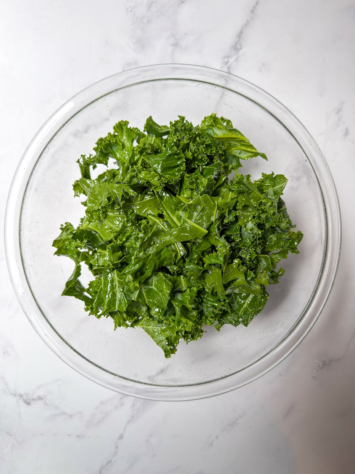 A clear glass bowl filled with fresh, chopped kale leaves sits on a white marble surface.