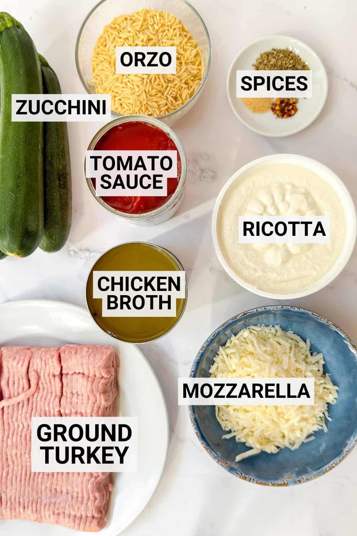 Labeled ingredients on a marble surface including zucchini, ground turkey, tomato sauce, chicken broth, ricotta, shredded mozzarella, and spices.