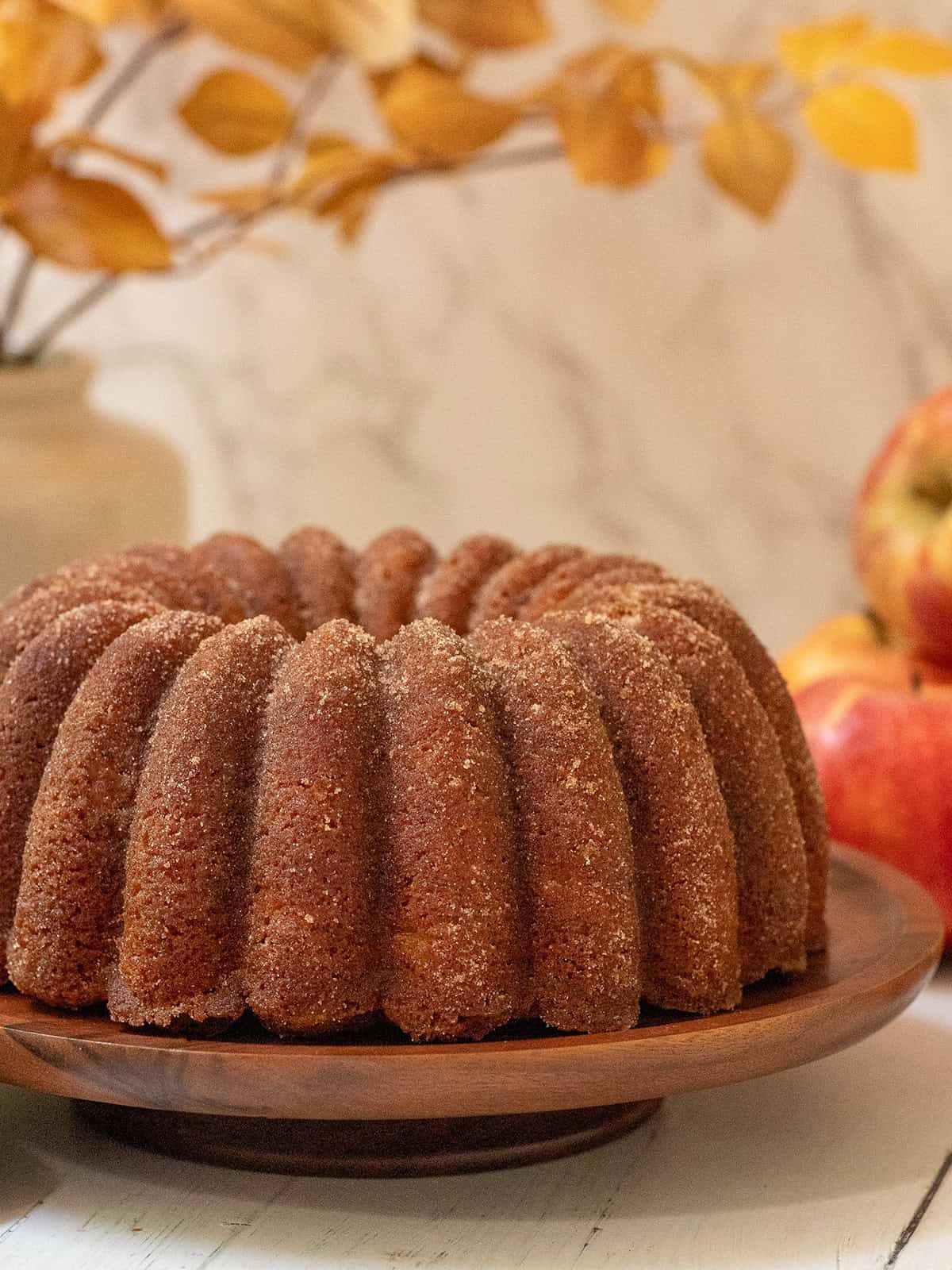 Cinnamon sugar apple cider Bundt cake on a wooden cake stand with apples and golden fall foliage in the background.