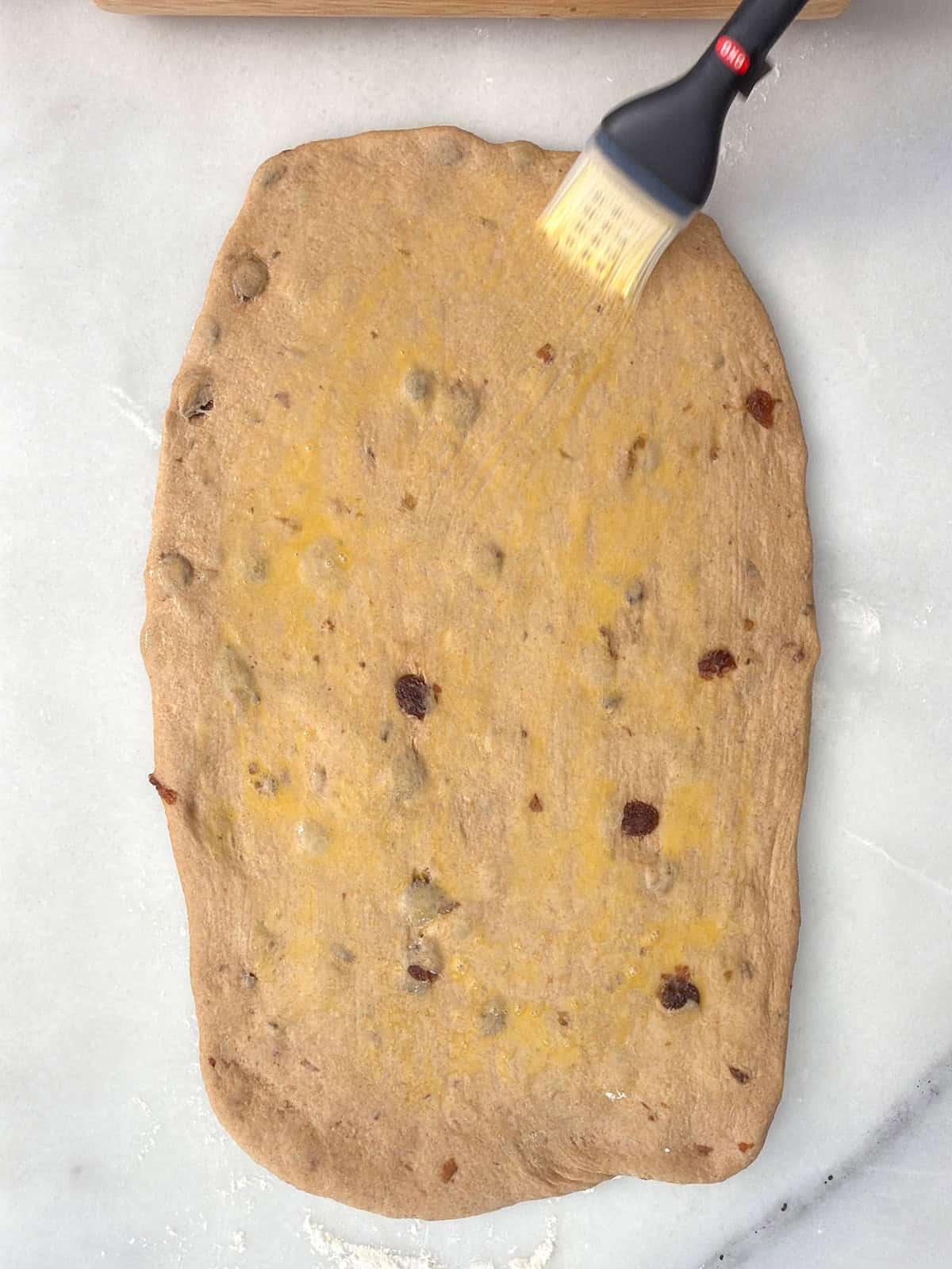 Brushing rolled whole wheat raisin bread dough with egg wash before adding cinnamon filling.