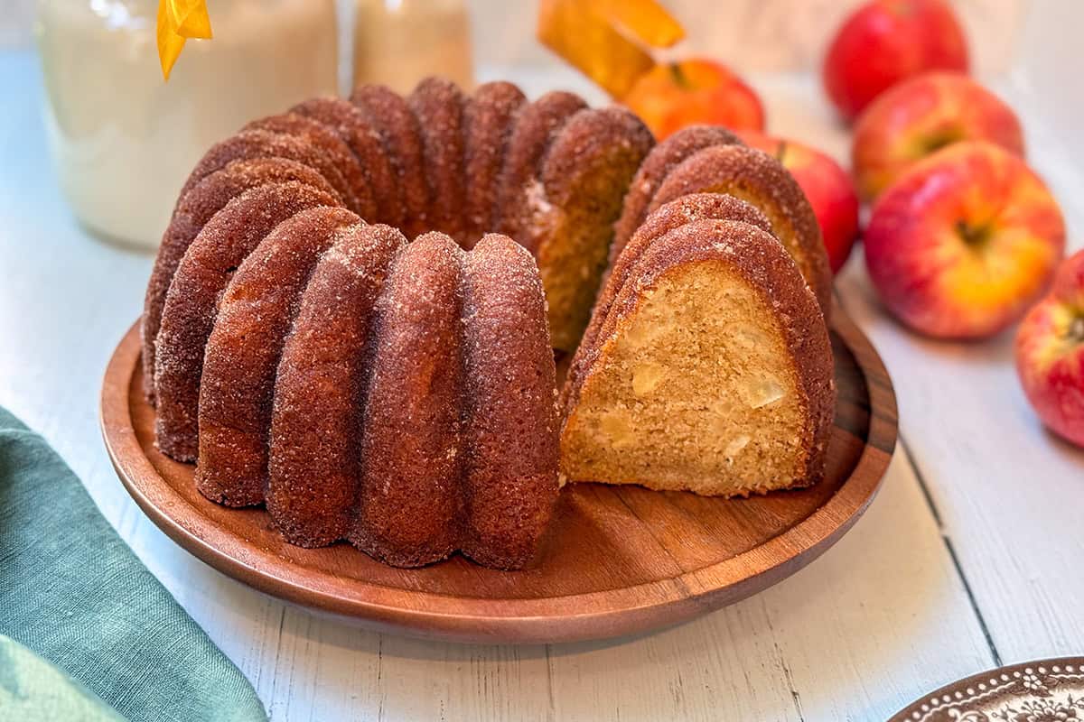 Apple cider donut Bundt cake with a slice cut to show the apple-filled crumb, surrounded by fresh apples and fall accents.
