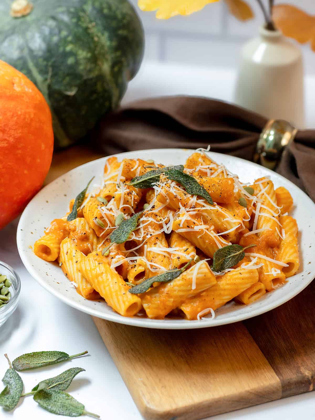 Creamy pumpkin sage pasta with rigatoni, crispy sage, Parmesan, black pepper, and pepitas on a white plate with fall pumpkins in the background.