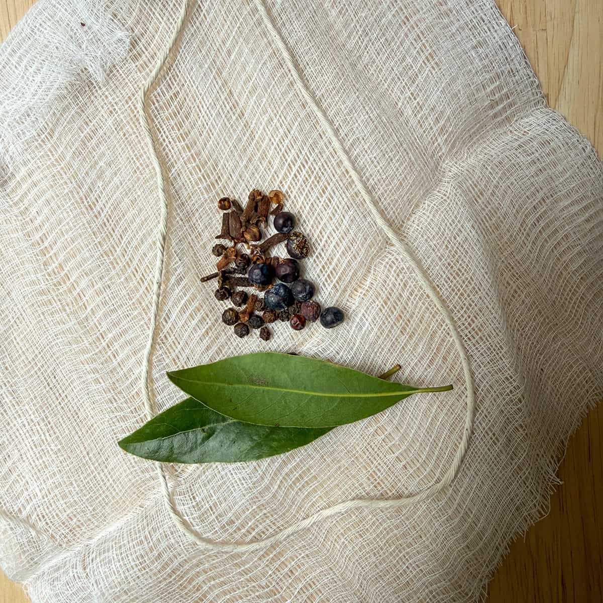 Sheet of cheesecloth with fresh bay leaves, whole cloves, and juniper berries with a kitchen twine to tie it into a bundle.