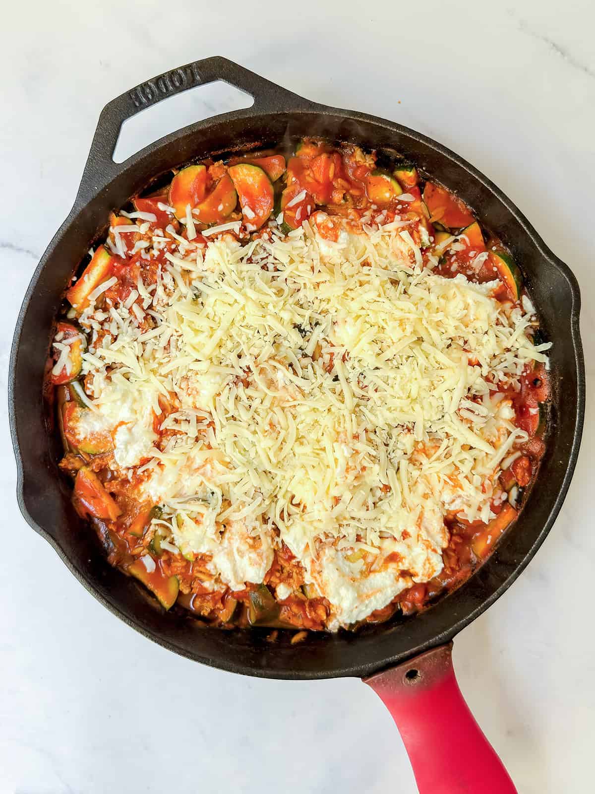Sprinkling grated mozzarella cheese over a cast iron skillet with ground turkey and zucchini in a creamy tomato sauce.