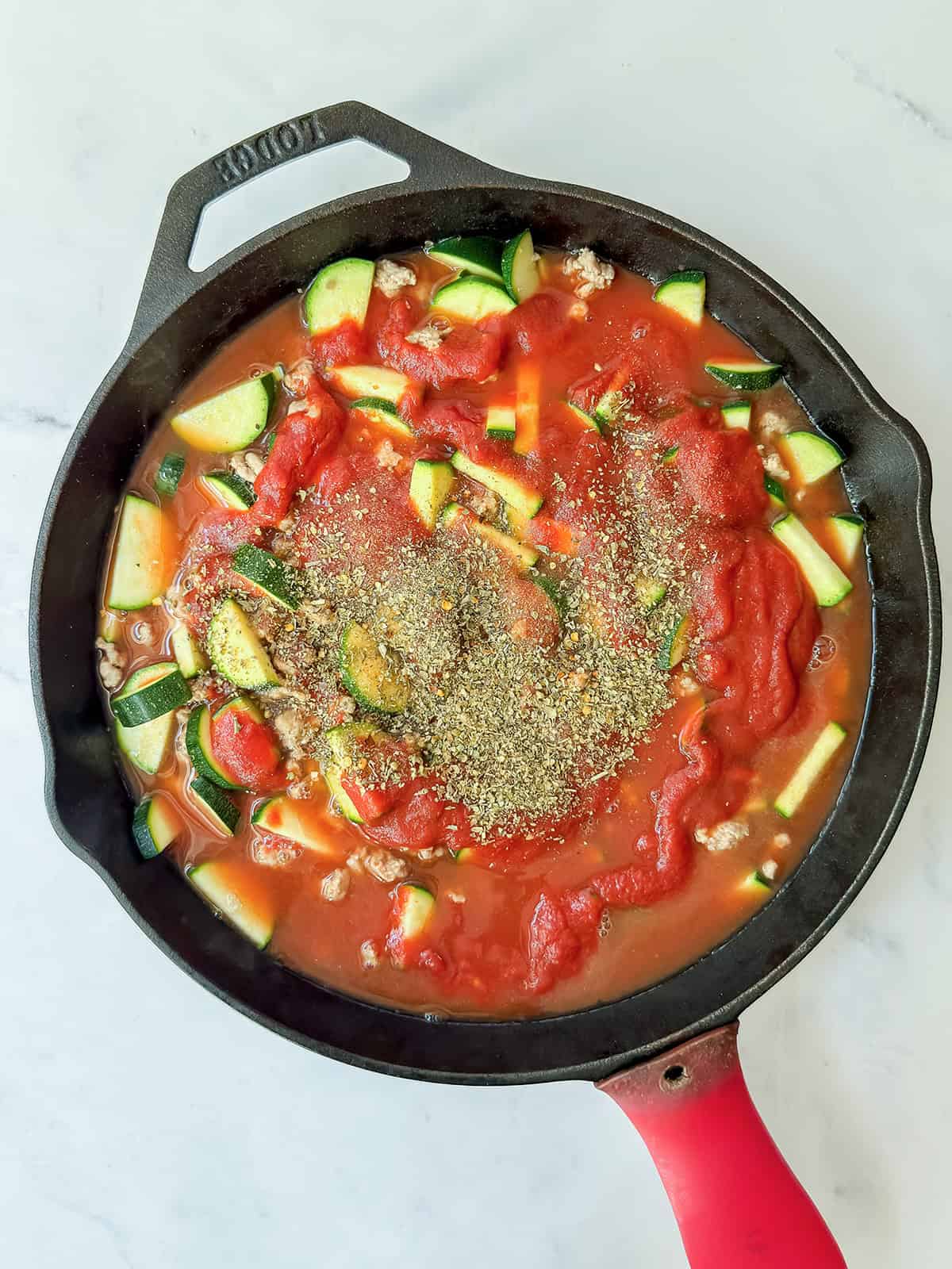 Adding tomato sauce and spices to a cast iron skillet with ground turkey and zucchini.