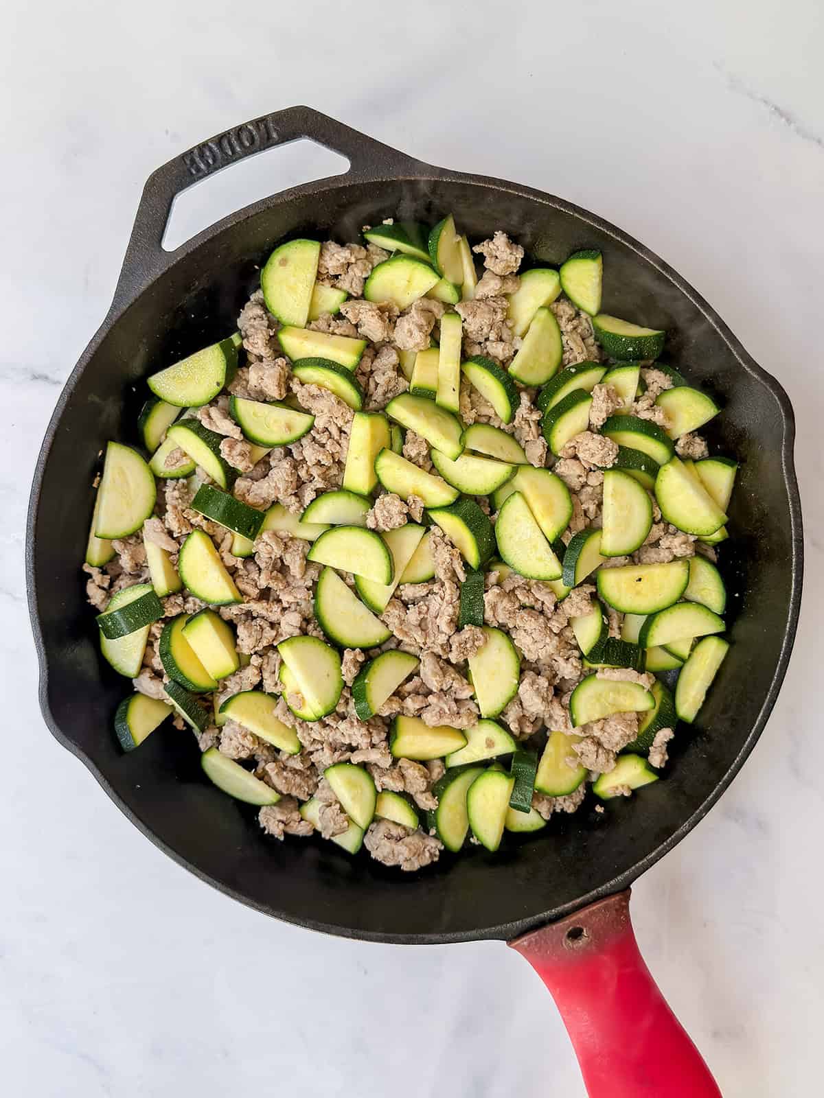Adding sliced zucchini to a cast iron skillet with browned ground turkey.